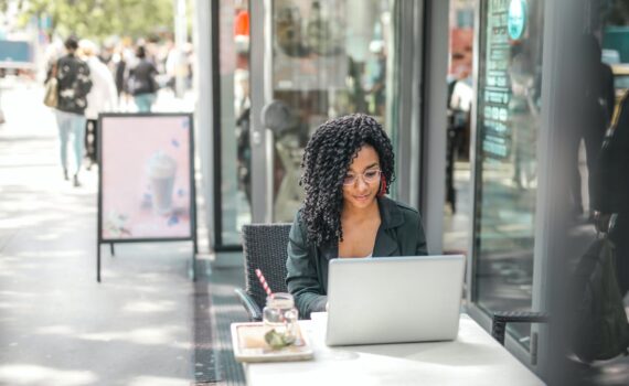 ethnic young woman using laptop while having tasty beverage in modern street cafe